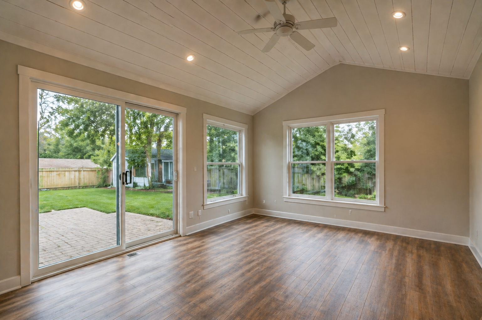 Spacious, empty living room before the sunroom addition. The room features wood-look flooring, neutral gray walls, recessed lighting, and a vaulted ceiling with a ceiling fan. Large sliding glass doors open to a backyard patio, showcasing a fenced yard and a small shed in the background.